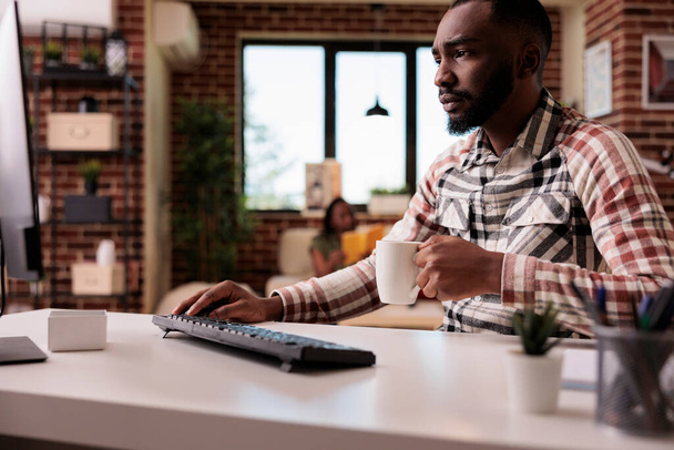 Zaměřený programátor držící šálek kávy nebo čaje vypadá ustaraně na obrazovce počítače, zatímco ladicí kód pracuje z domova. Freelancer čtení týkající se e-mailu na PC, zatímco sedí u stolu. - Fotografie, Obrázek