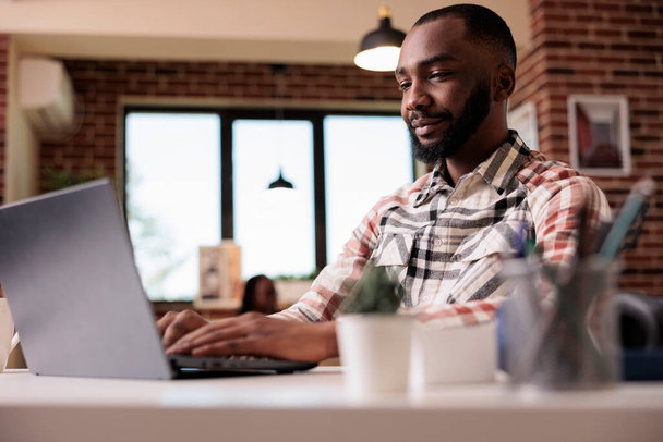 Portré afro-amerikai szabadúszó dolgozik távoli gépelés és nézi laptop képernyőn, miközben barátnő pihen a kanapén. Férfi programozó segítségével hordozható számítógép fejleszteni szoftver otthon. - Fotó, kép