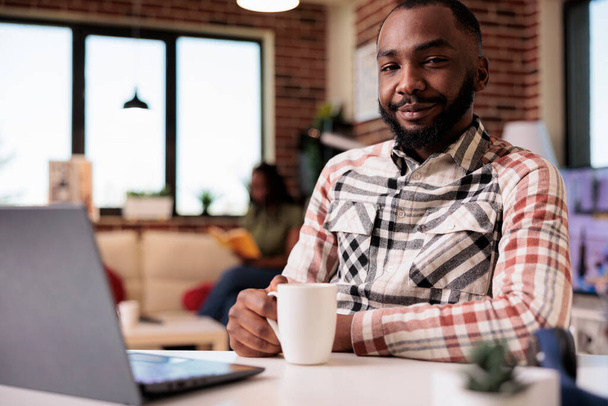 African american programmer posing confident looking at camera holding a cup of coffee while girlfriend is reading a book. Freelancer coder taking a break relaxing watching video content on laptop. - Fotó, kép
