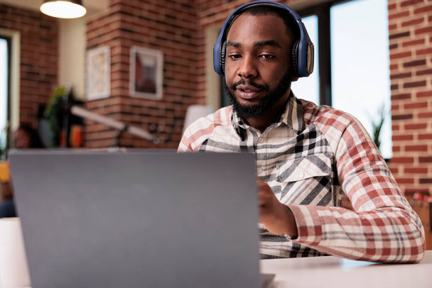 リビングルームのデスクに座っている間、ラップトップでクライアントに耳を傾けながら、ビデオ会議でワイヤレスヘッドフォンを持つフリーランサー。アフリカ系アメリカ人の起業家はインターネットコールに焦点を当て. - 写真・画像