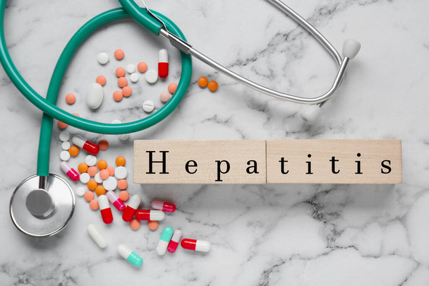 Слово Гепатит сделан из деревянных кубиков, стетоскоп и таблетки на белом мраморном столе, плоский лежал - Фото, изображение