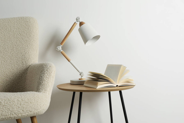 Stylová lampa s otevřenou knihou na dřevěném konferenčním stolku a měkkém křesle u bílé stěny. Návrh interiéru - Fotografie, Obrázek