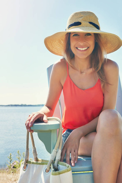 Im going to stay a bit longer. Portrait of a cheerful young woman wearing a hat while being seated on a chair next to a lake outside in the sun - Photo, image