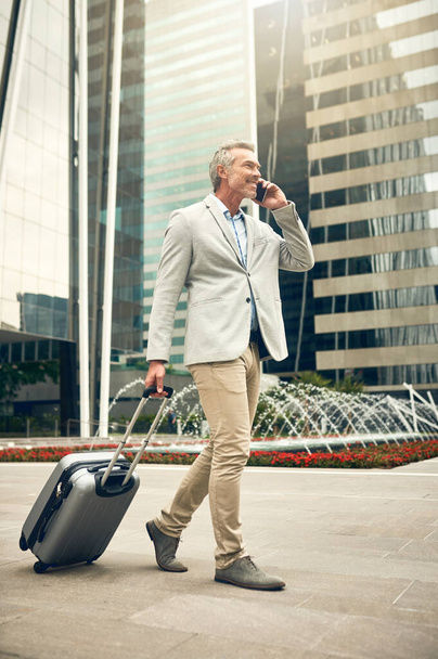 Bonjour le succès, je suis arrivé. un homme d'affaires mature parlant sur un téléphone portable tout en marchant avec une valise dans la ville - Photo, image