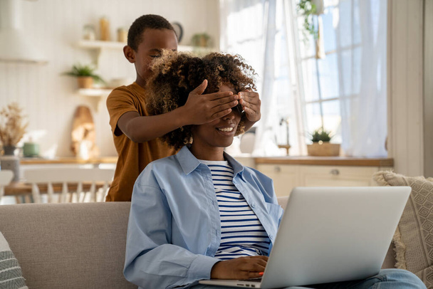 Smiling little African American boy son covering eyes of mother freelancer at home, distracting mom from remote job, asking for attention. Woman mom sitting on couch trying to work on laptop with kid - Photo, Image