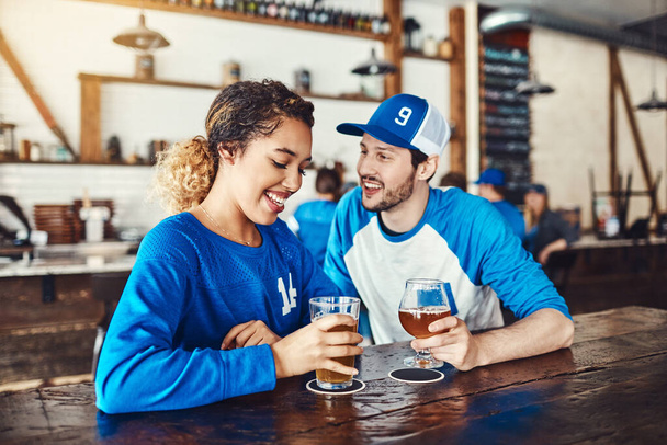 Είσαι κι εσύ φίλαθλος. ένας νεαρός άνδρας και μια γυναίκα που πίνουν μπύρες ενώ βλέπουν ένα αθλητικό παιχνίδι σε ένα μπαρ - Φωτογραφία, εικόνα