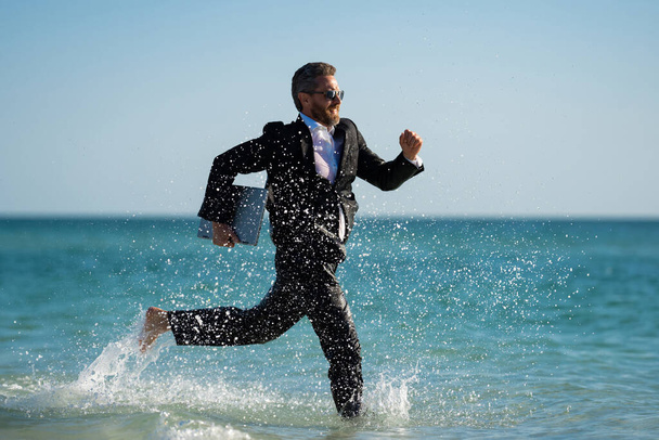 Business man in suit with laptop running in sea water. Travel tourism and business concept. Crazy male office employee with laptop running in sea. Crazy business man on summer vacation - Fotoğraf, Görsel