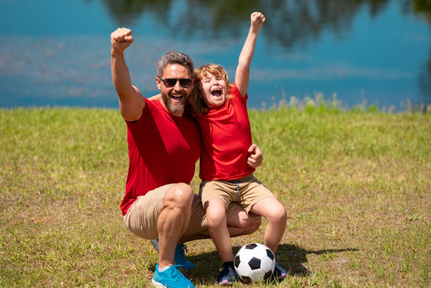 Dad with his little cute son are having fun and playing football on green grassy summer lawn. Football soccer sport concept. Father and son celebrate together after scoring goal or making a good play - Fotoğraf, Görsel
