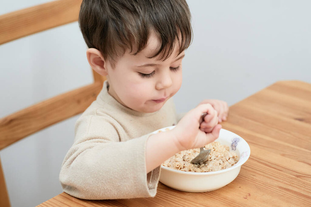 маленький мальчик ест овсянку на завтрак на кухне - Фото, изображение
