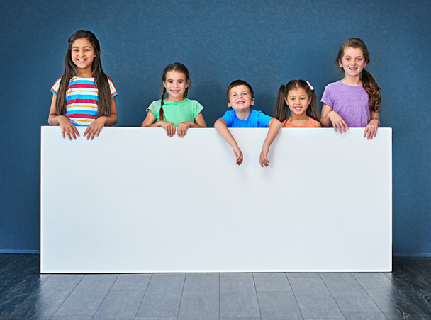 Tenemos un mensaje para ti. Captura de estudio de un grupo diverso de niños de pie detrás de una gran pancarta en blanco sobre un fondo azul - Foto, imagen