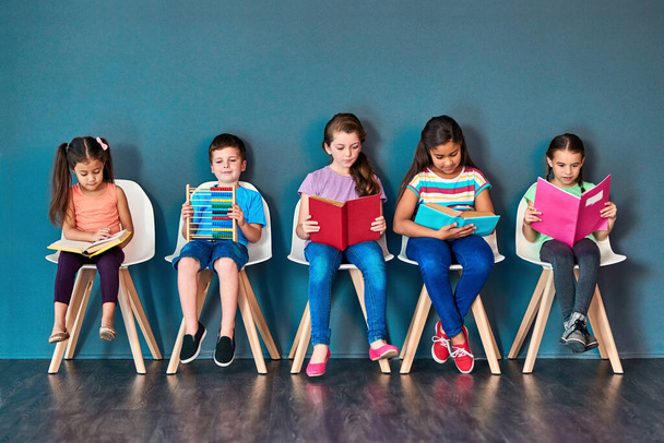 Багато чому навчитися. Студійний знімок групи дітей, які сидять на стільцях і читають книги на синьому фоні
 - Фото, зображення