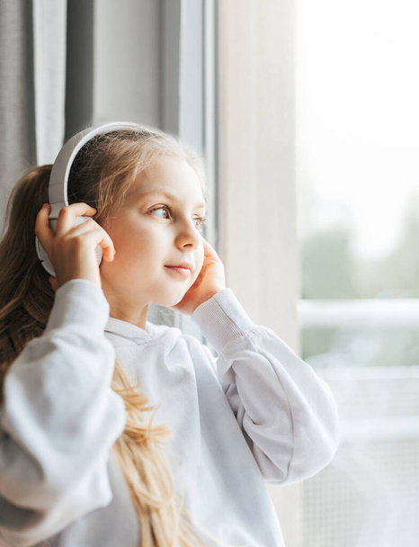 Little girl with headphones indoors at home, sitting on window sill and listening music - Photo, Image