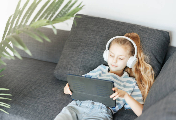 Niña acostada en el sofá con una tableta electrónica escucha música en auriculares blancos. Uso de dispositivos inalámbricos para la educación. - Foto, imagen