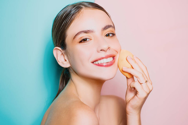 Healthy smiling girl with bare shoulders, clear skin, dark hair and beige sponge. Natural makeup look. Health and beauty concept. Happy young woman covering her skin with foundation - Photo, image