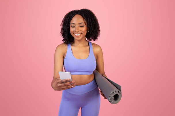 Smiling young african american lady in sportswear hold mat, chatting in smartphone, enjoy sports alone on pink studio background. Fit lifestyle, body care, app for workout, social networks - Фото, изображение