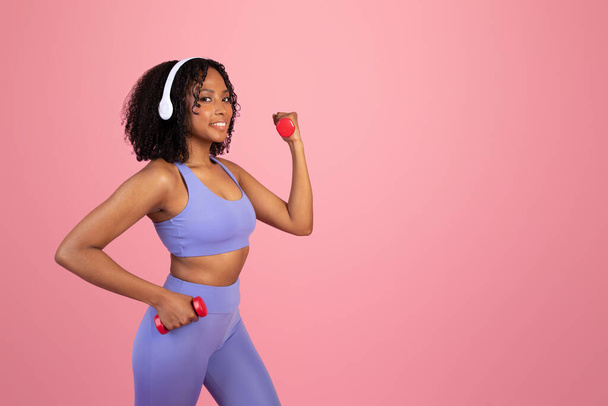 Happy young black woman in sportswear and wireless headphones doing hand exercise with dumbbells, enjoy sport with music alone on pink studio background. Fit lifestyle, body care and workout - Foto, afbeelding