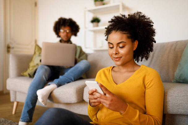 Siyah kadın akıllı telefonda sörf yaparken kocası evdeki koltukta dizüstü bilgisayar kullanıyordu. Hafta sonları Modern Oturma Odasında Gadget kullanan genç bir çift. Seçici Odaklanma - Fotoğraf, Görsel