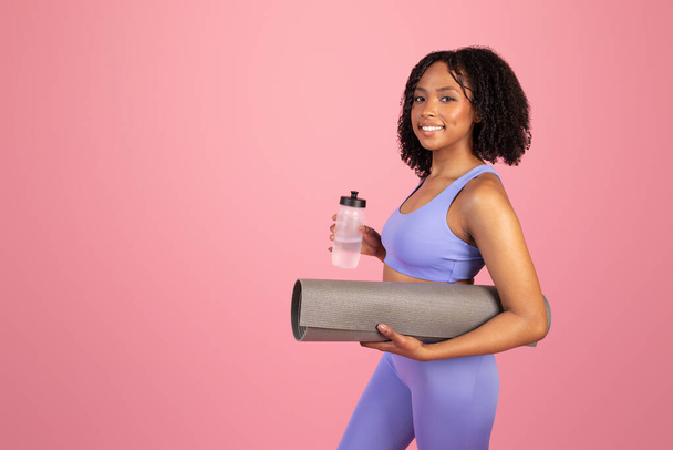 Frohe junge afrikanisch-amerikanische Lockenkönigin in Sportbekleidung mit Matte und Flasche Wasser genießt Sport allein auf rosa Studiohintergrund. Empfehlung Fit Lifestyle, Körperpflege und Workout - Foto, Bild