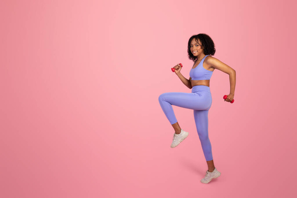 Веселая молодая африканская американка в спортивной одежде делает кардио-упражнения с гантелями для рук, наслаждается спортом в одиночестве на розовом фоне студии, полной длины. Подходящий образ жизни, реклама и предложение, уход за телом - Фото, изображение