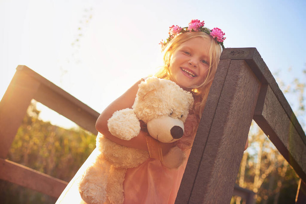 Shes having a great day with best friend. a happy little girl holding a teddy bear and looking at the camera while standing on a bridge - Photo, Image