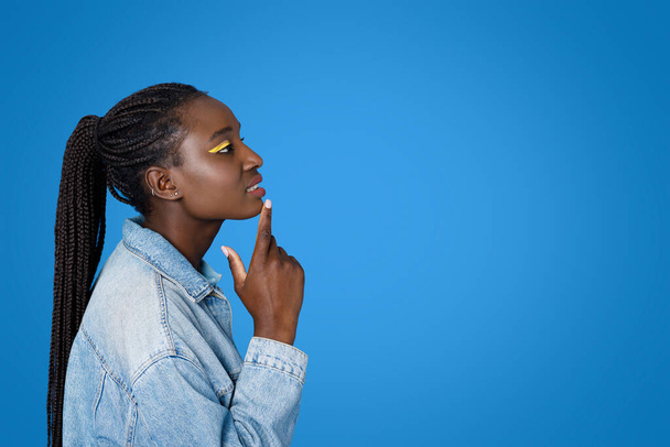 Ciekawy atrakcyjny stylowy młody czarny kobieta w casual strój i jasne oczy dotyka podbródek i patrząc na przestrzeni kopii, izolowane na niebieskim tle studio, widok z boku - Zdjęcie, obraz