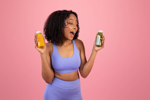 Onnellinen nauraa nuori afrikkalainen amerikkalainen kihara nainen urheiluvaatteet osoittavat smoothie tai mehua pulloja, eristetty vaaleanpunainen studio tausta. Sopiva, aktiivinen elämäntapa, kehonhoito, ruokavalio ja terveellinen juoma - Valokuva, kuva