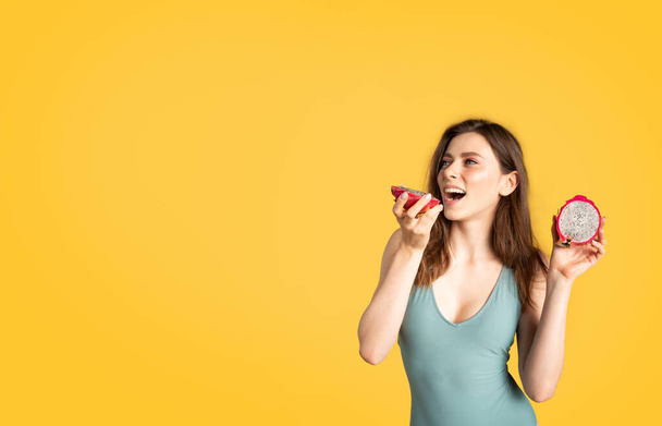 Joyful young lady in swimsuit biting pitaya slice, posing with dragon fruit and enjoying tasty juicy fruit, standing on yellow studio background with free space - Photo, Image
