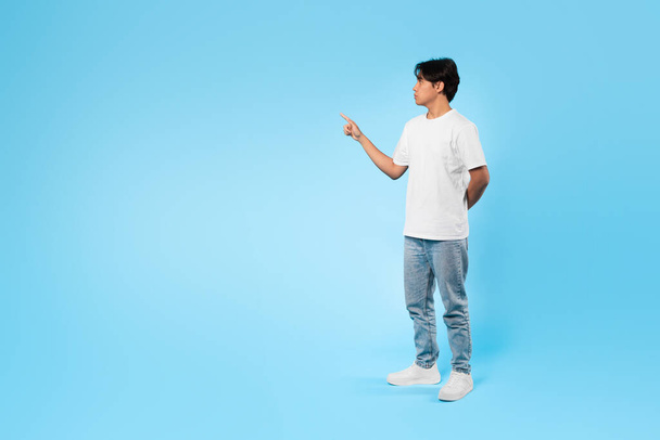 Look Aside. Asian Student Teen Guy Pointing Finger At Free Space Advertising Text Offer Standing Over Blue Studio Background. Full Length Shot Of Boy Pushing Invisible Button. Side View - Photo, image