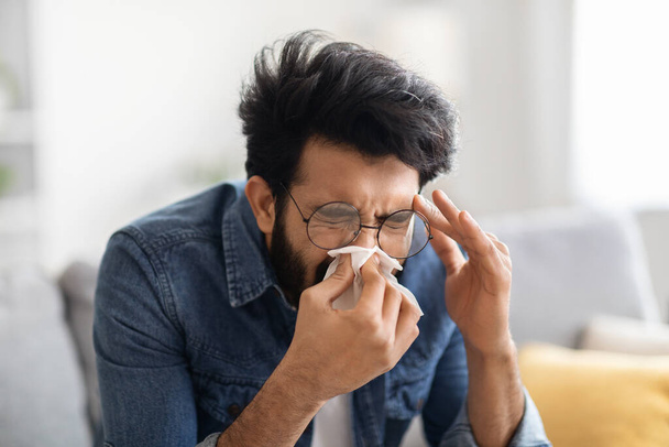 自宅でソファに座っている間、紙のティッシュで鼻を吹いて病気の若いインド人男性,季節のインフルエンザや風邪を患っている千年紀の東の男,鼻炎やアレルギー反応を持っています,閉鎖ショット - 写真・画像