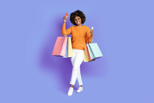 Δροσερό κομψό χαρούμενο χαμόγελο νεαρή μαύρη γυναίκα με φουντωτά μαλλιά φορώντας γυαλιά ηλίου κρατώντας πολύχρωμες τσάντες αγορών, smartphone, τραπεζική κάρτα, θέτοντας πάνω από μωβ φόντο στούντιο. Απεριόριστα ψώνια - Φωτογραφία, εικόνα