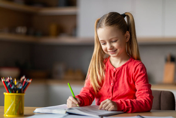 Portrait de mignonne petite fille faisant des devoirs scolaires avec livre à la maison, souriant Preteen fille assise au bureau dans la cuisine et écrivant des notes dans le classeur, appréciant l'étude, l'espace de copie - Photo, image