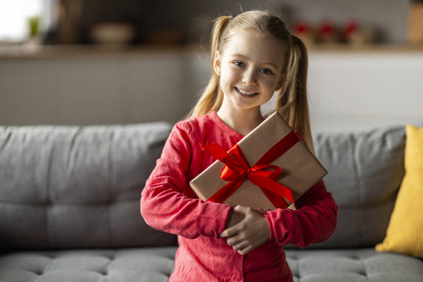 Милая маленькая девочка позирует с подарочной коробкой в руках в интерьере дома, счастливая Preteen ребенок держит завернутый подарок с красной лентой и улыбаясь на камеру, празднуя день рождения, копировать пространство - Фото, изображение