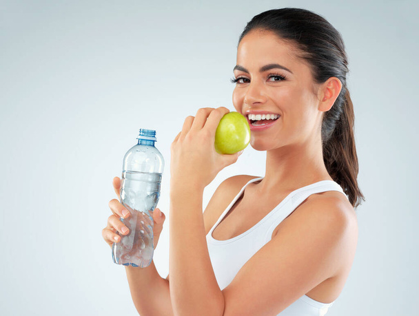 Eat healthy. Your body is worth it. Studio shot of a fit young woman holding a bottle of water and an apple against a gray background - Фото, изображение