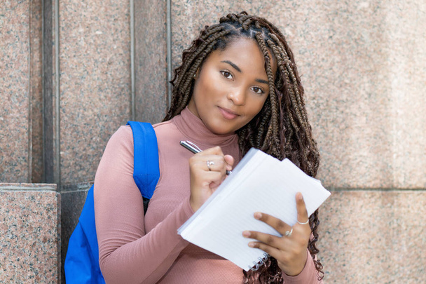 African american female university student with backpack writing notess outdoors in city in summer - Φωτογραφία, εικόνα