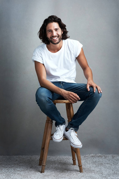 His smile is part of his charm. Studio shot of a handsome young man sitting on a stool against a grey background - Photo, Image