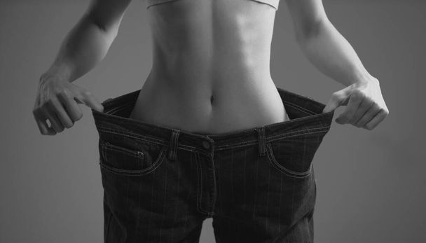 Skinny waist. Slim girl have points at slim waist in big trousers, successful weight loss, isolated on gray background. Successful weight loss, woman with too large jeans after a diet - 写真・画像