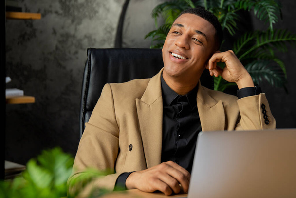 Smiling Black businessman in jacket using laptop in cozy office with plants. Successful African American entrepreneur enjoying work day. High quality photo - Photo, Image