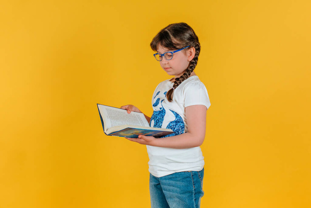 smart beautiful girl with pigtails over isolated background holding a book in her hand. Schoolgirl with books and glasses on a yellow background. - Photo, image
