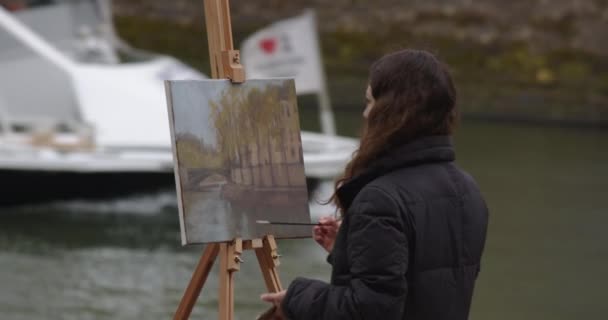 Een vrouw die een schilderij schildert bij de Seine. Creatieve kunstenares schildert een schilderij met een borstel terwijl ze bij de ezel in Parijs staat. Vrouw kunstenaar, kunst voor verkoop, inspiratie. - Video