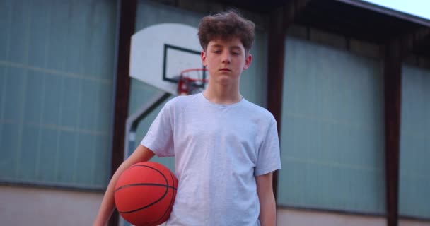 Video van een jongen die op een basketbalveld staat en een basketbal vasthoudt. Cinematografisch schot van tiener staan met een bal in zijn hand en een basketbal mand achter hem. - Video