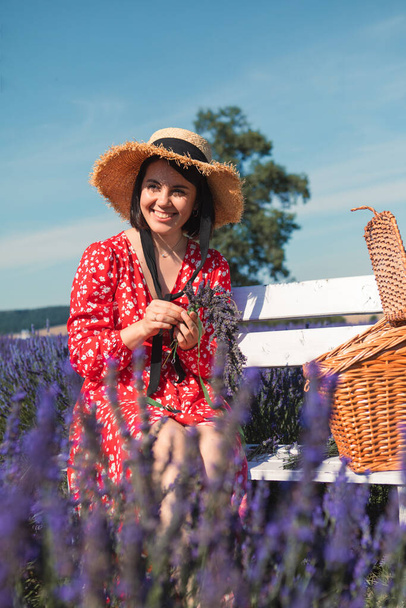 a young woman in a straw hat sits on white bench in a lavender field and weaves a wreath - Foto, imagen