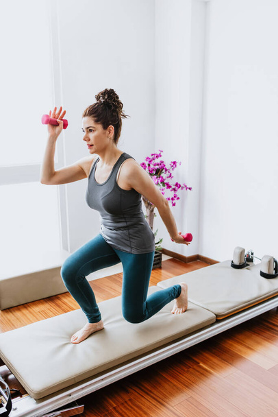 latin adult woman doing pilates exercises workout on reformer bed at home in Mexico, hispanic people in Latin America - Photo, Image