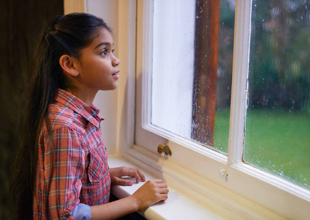 Rain, window and girl with ideas, thinking or wonder with thoughts, ponder or kid at home. Female person, thoughtful child and model with winter weather, house and storm with fantasy and steamy glass. - Photo, Image