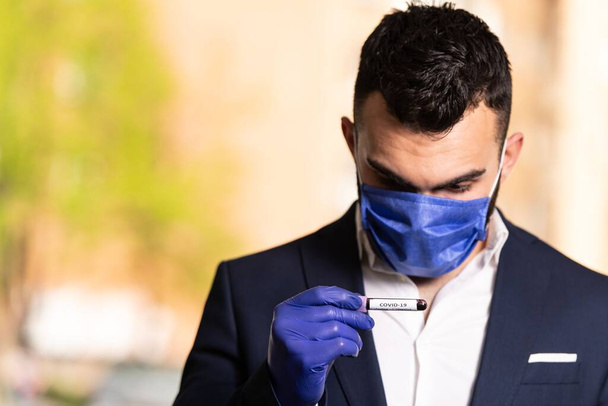 A handsome businessman with a surgical mask and gloves holding a blood test tube in a park - Fotoğraf, Görsel