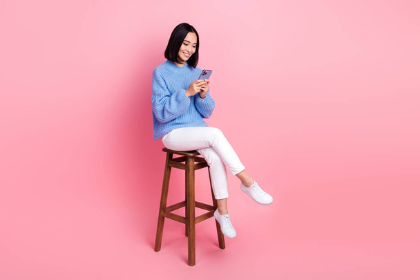 Full length photo of cheerful positive girl wear blue sweater white pants sitting on chair read message isolated on pink color background. - Photo, image