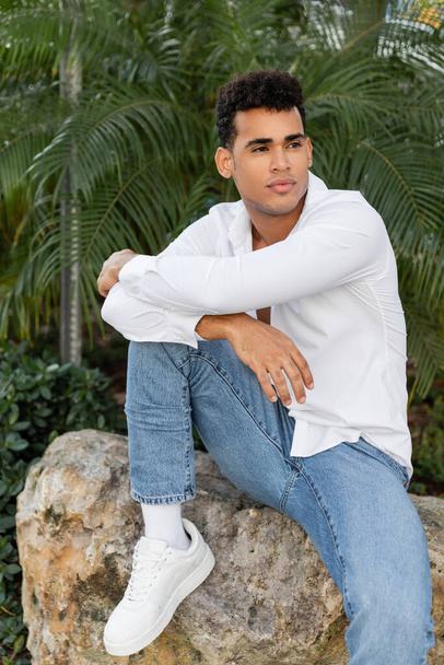 Joven y elegante hombre cubano con camisa blanca y jeans mirando hacia otro lado mientras está sentado en piedra en Miami - Foto, Imagen