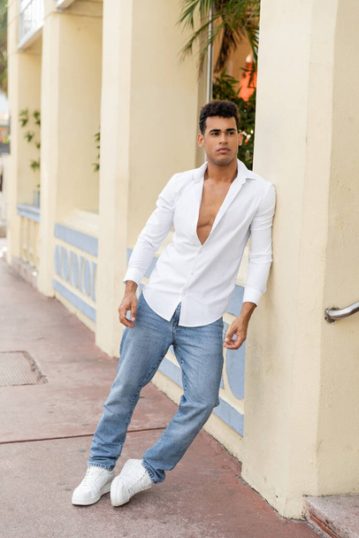 hombre cubano bien parecido en camisa y jeans mirando hacia otro lado cerca de edificio en la calle urbana en Miami - Foto, imagen