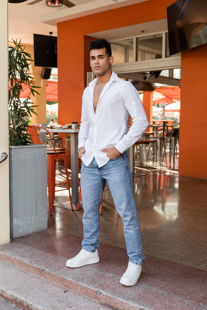 Pełna długość Kubańczyka w białej koszuli i dżinsach pozujących w kawiarni na ulicy miejskiej w Miami - Zdjęcie, obraz