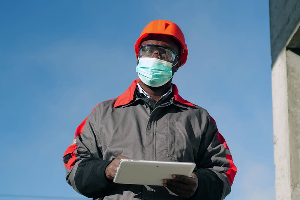 Afrikkalainen amerikkalainen työntekijä lääketieteellisessä kasvonaamiossa seisoo rakennustyömaalla tabletin kanssa. Workman omistaa käsissä valkoinen tabletti PC ja ajattelee - Valokuva, kuva