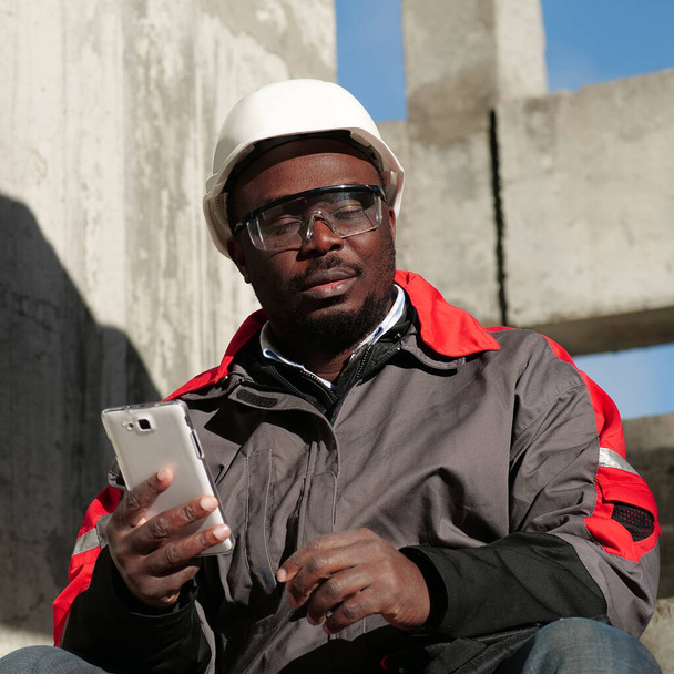 Κουρασμένος εργάτης κάθεται στις σκάλες με κλειστά μάτια και κρατά σε χέρια λευκό smartphone. Αφροαμερικανός εργάτης με σκληρό καπέλο και προστατευτικά γυαλιά με smartphone κάθεται στο εργοτάξιο - Φωτογραφία, εικόνα